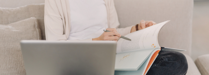 Une femme révise sur son canapé à l'aide d'un livre et de son ordinateur portable.